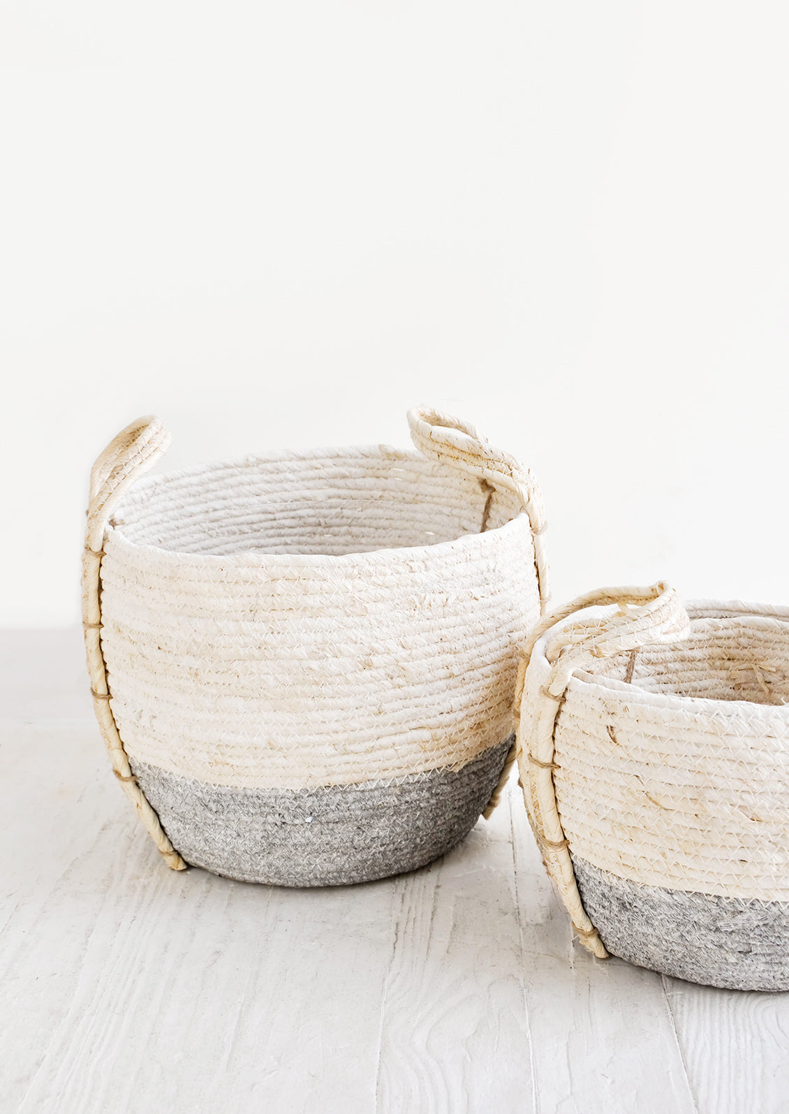 Small, round storage baskets made from natural maize fiber, fiber handles attached at sides, band of contrasting grey color along bottom.