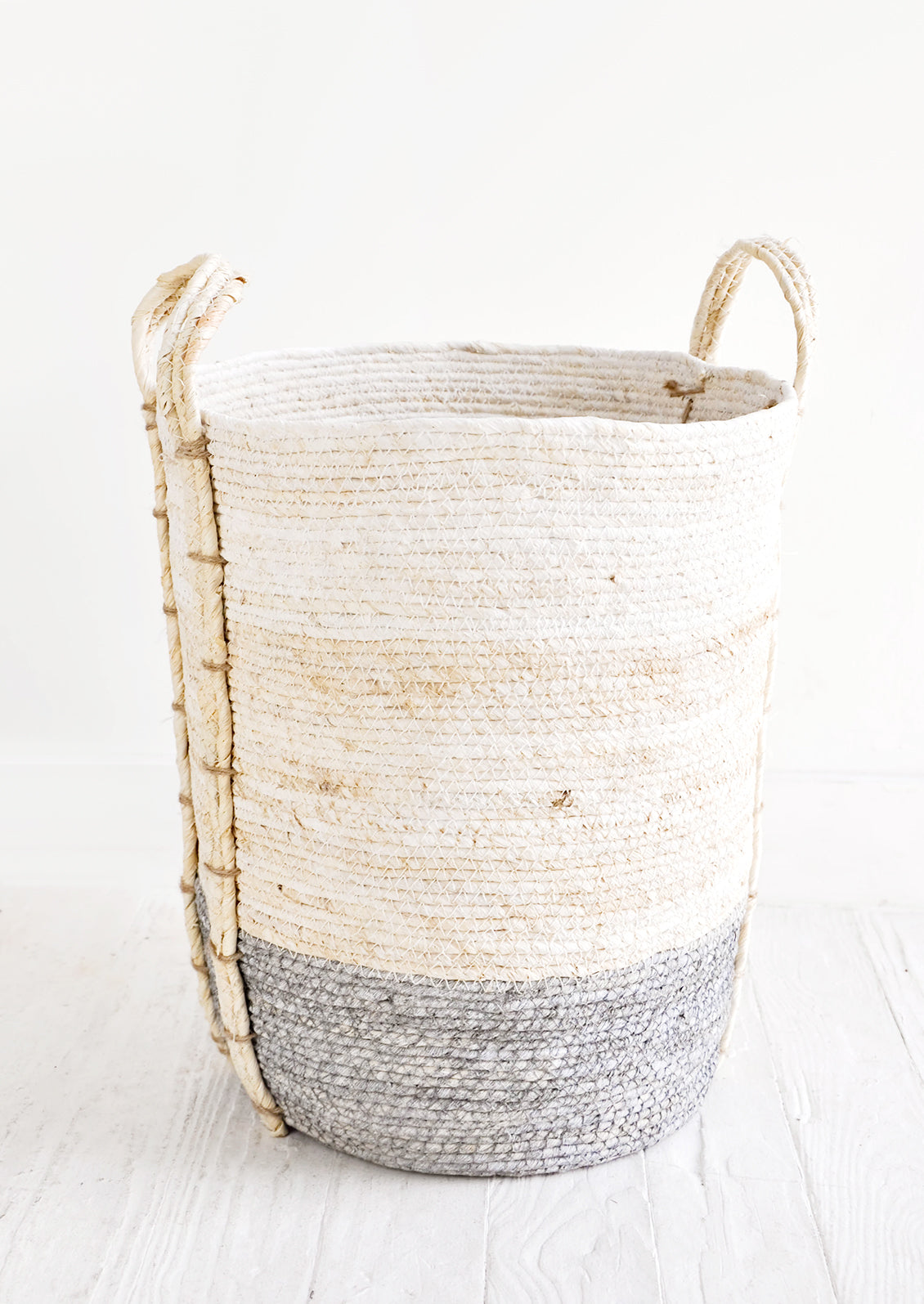 Round, tall storage basket made from natural maize fiber, fiber handles attached at sides, band of contrasting grey color along bottom.