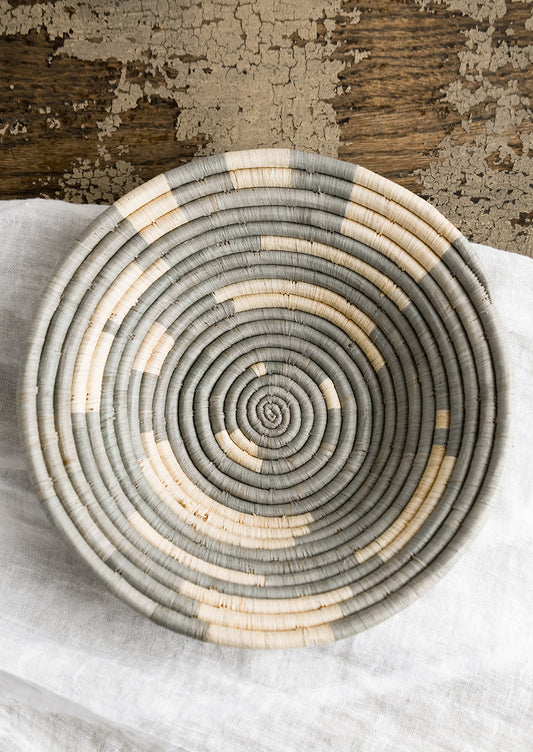 A round raffia bowl in dusty blue-grey and natural spiral geometric pattern.