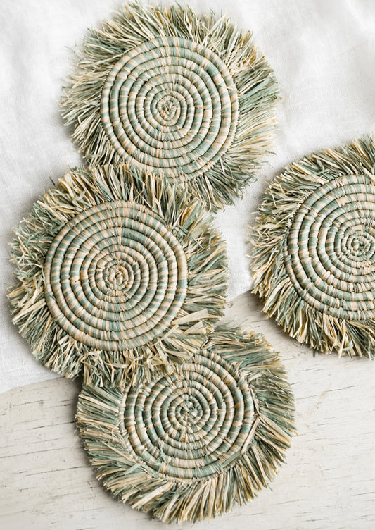 A set of four natural raffia coasters with heathered seafoam color and fringe trim.