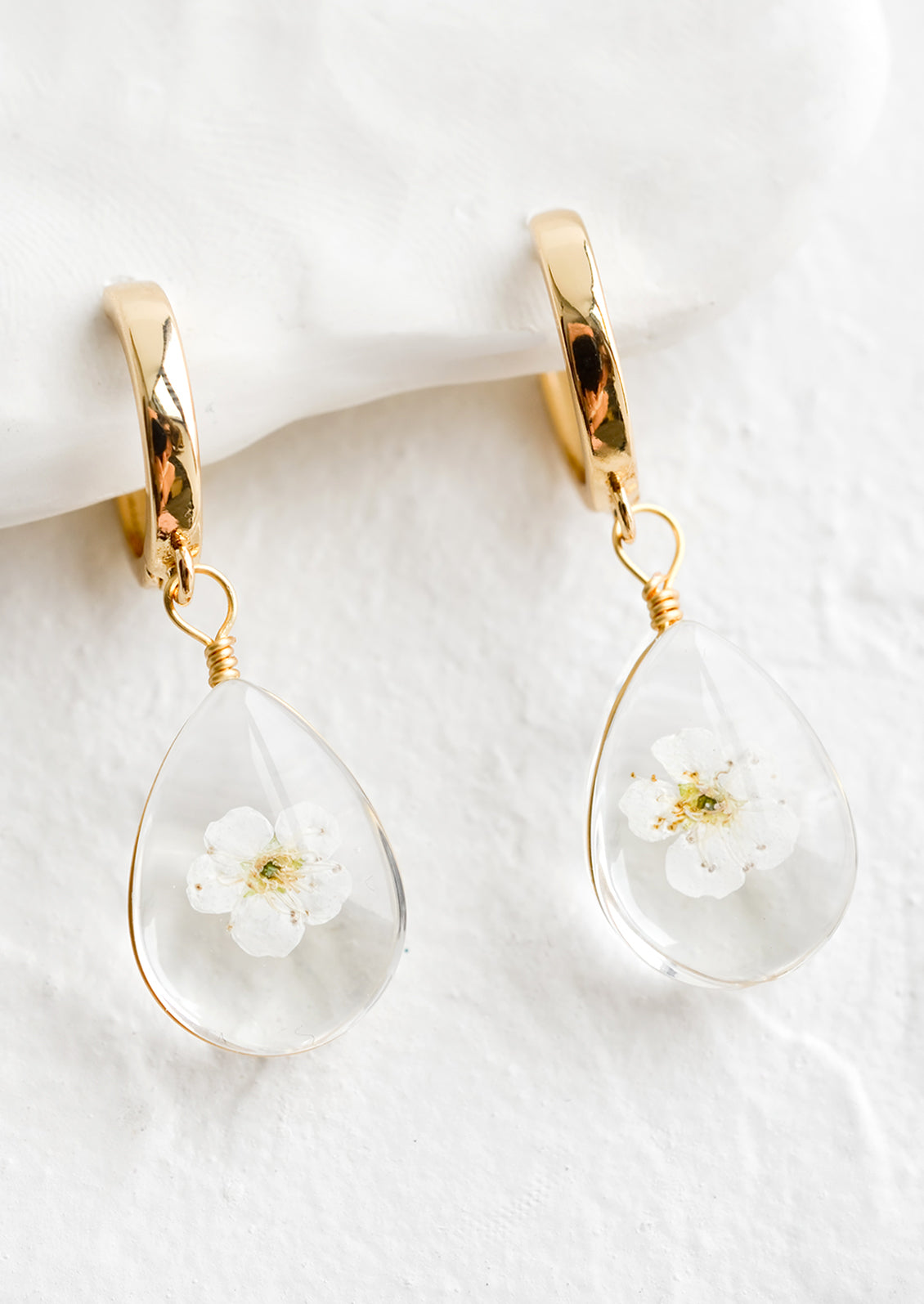 A pair of earrings with gold huggie hoop and clear resin drop with encased white florals.