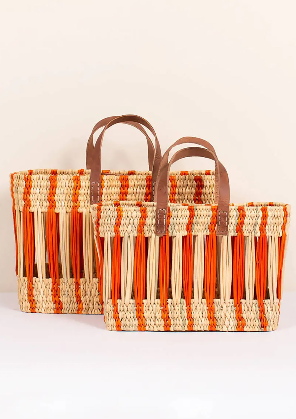 An oblong open weave basket in natural reed with bright orange stripes and brown leather handles.