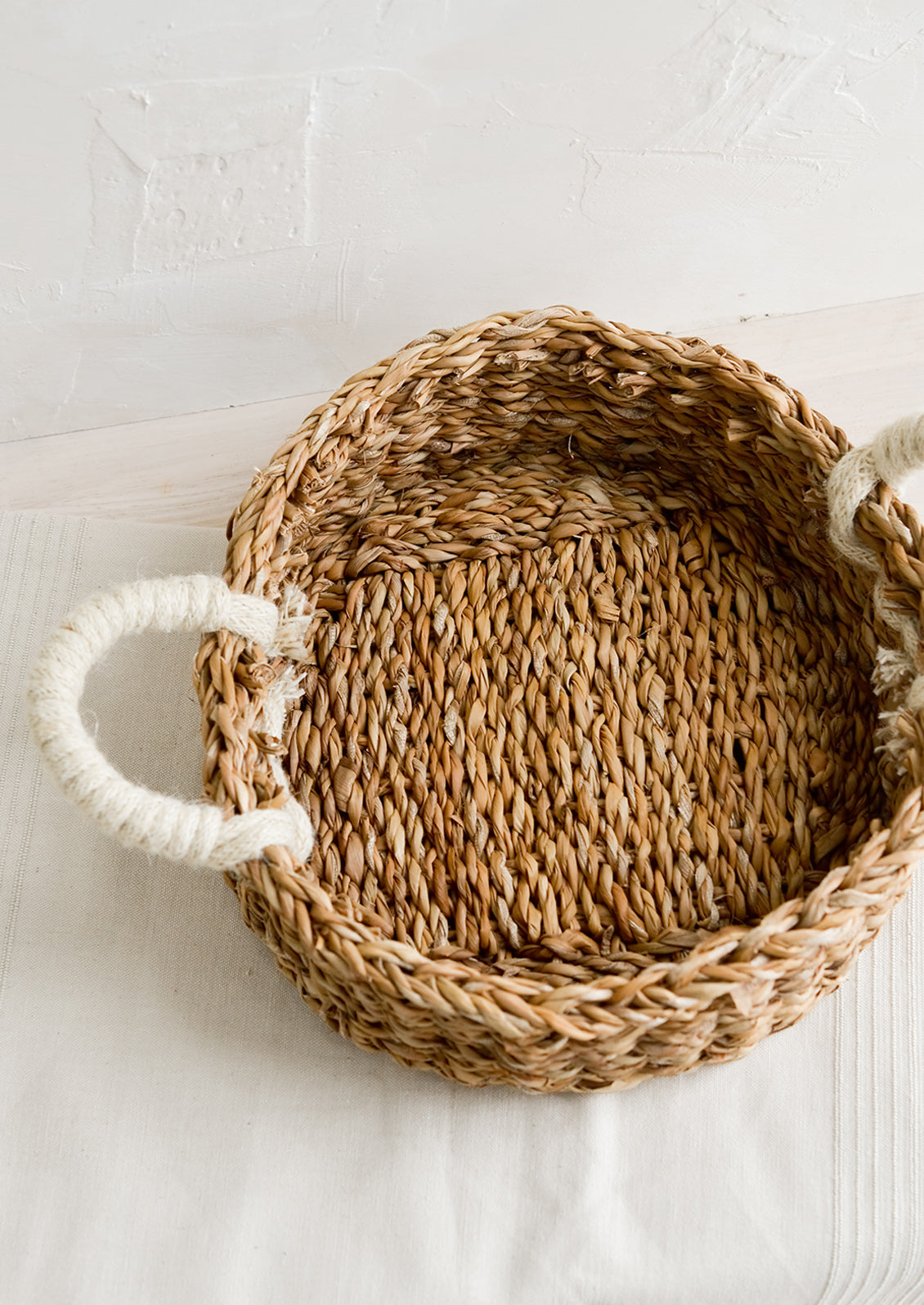 A round, shallow tray made from seagrass with ivory jute handles at sides.