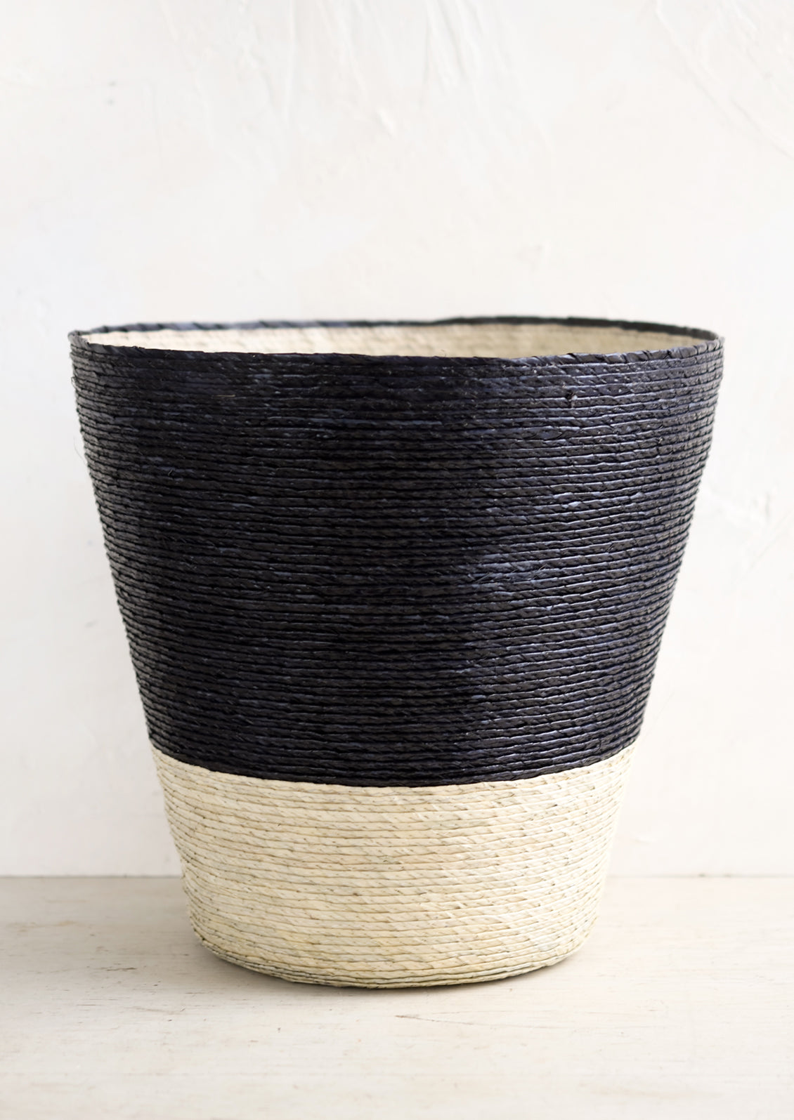 A conical shaped storage basket made from woven palm leaf in black & natural color way.