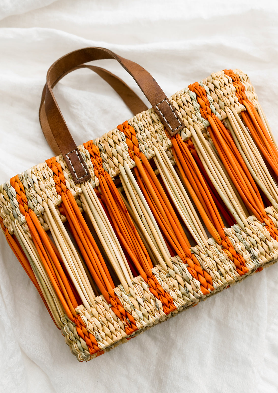 An oblong open weave basket in natural reed with bright orange stripes and brown leather handles.