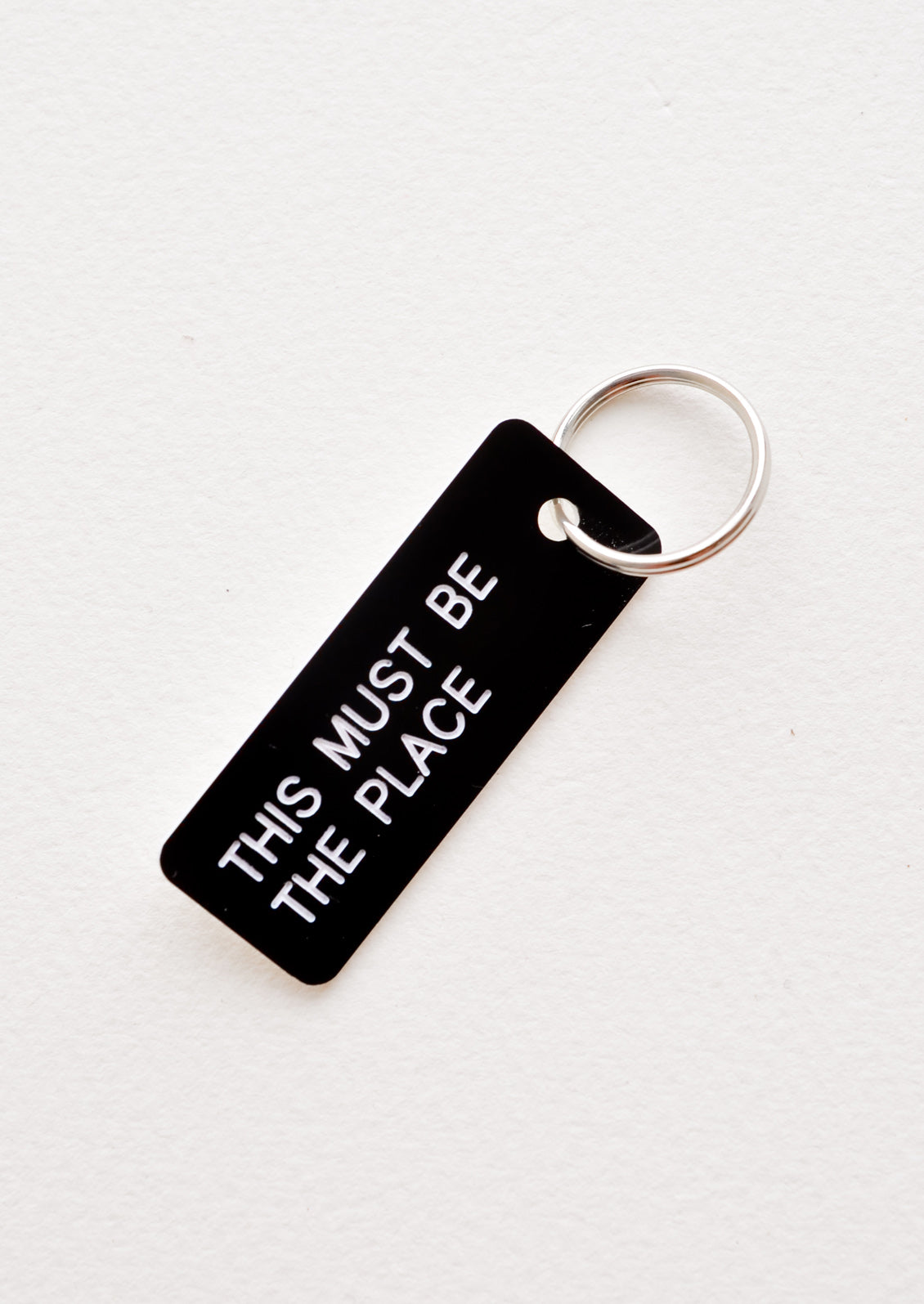 Small acrylic keychain, black background with white words that says "THIS MUST BE THE PLACE"