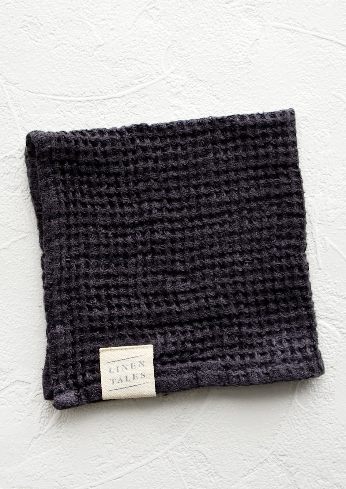 A folded waffle-weave linen washcloth in charcoal with a logo patch.