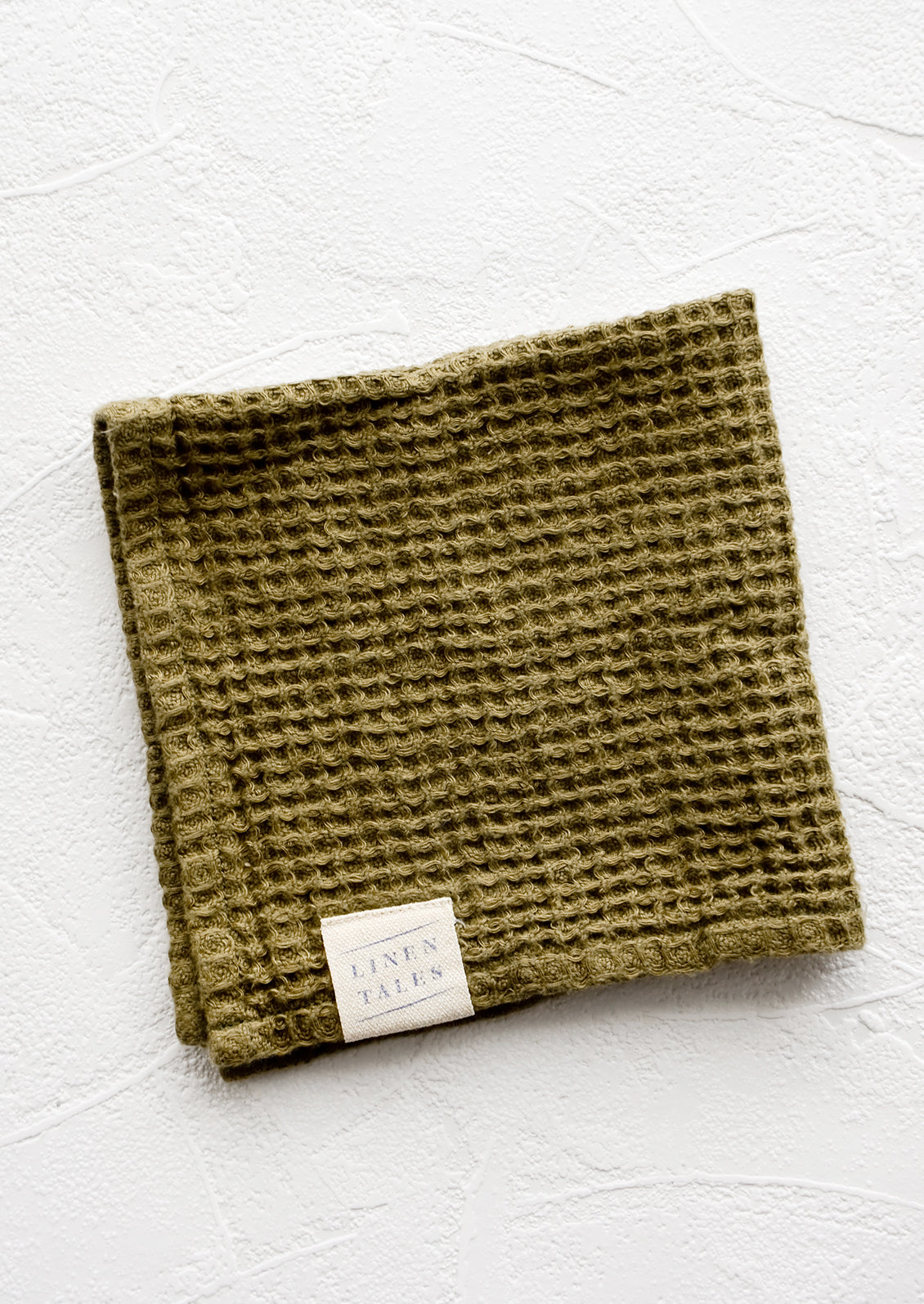 A folded waffle-weave linen washcloth in olive green with a logo patch.