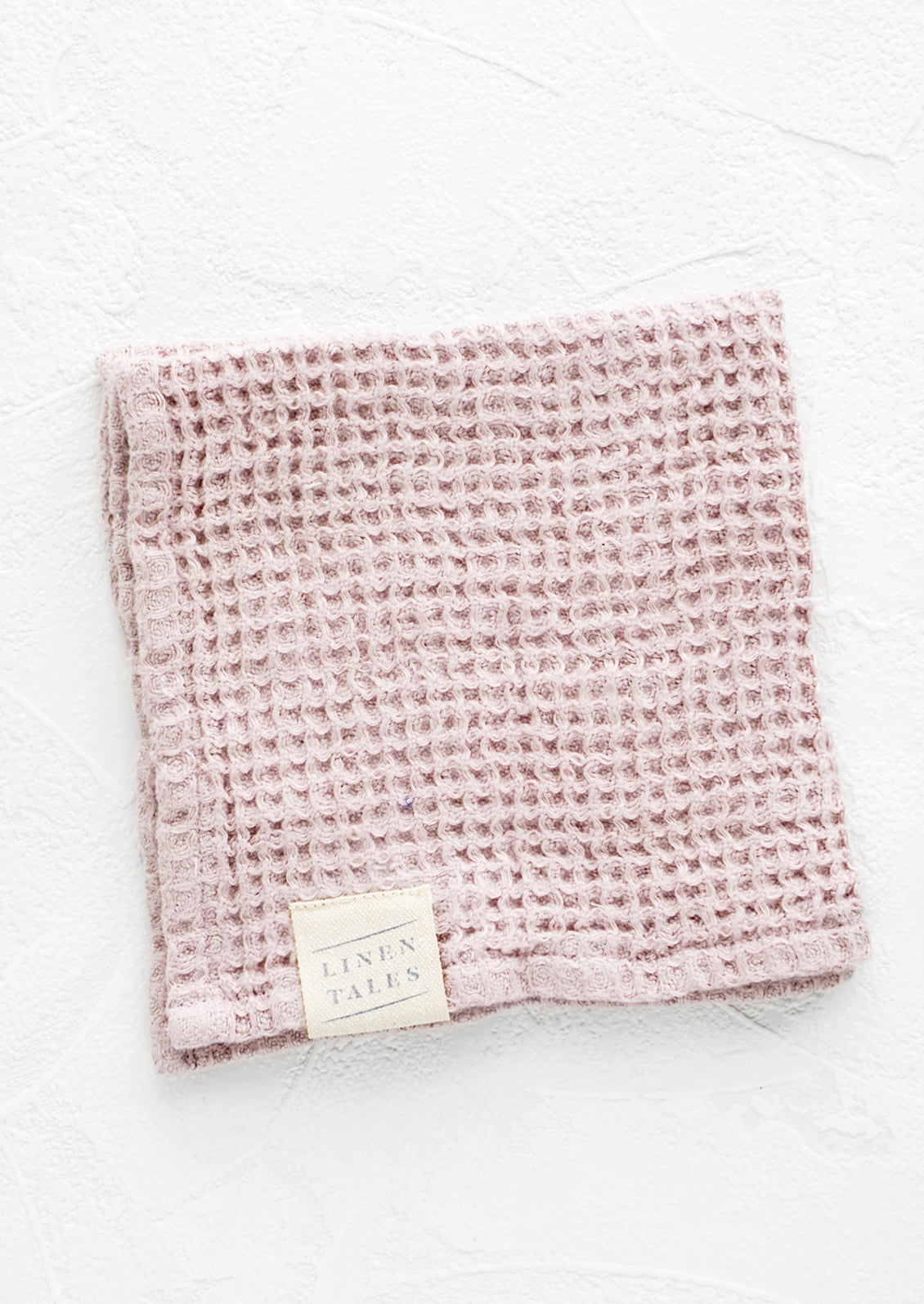 A folded waffle-weave linen washcloth in pale pink with a logo patch.