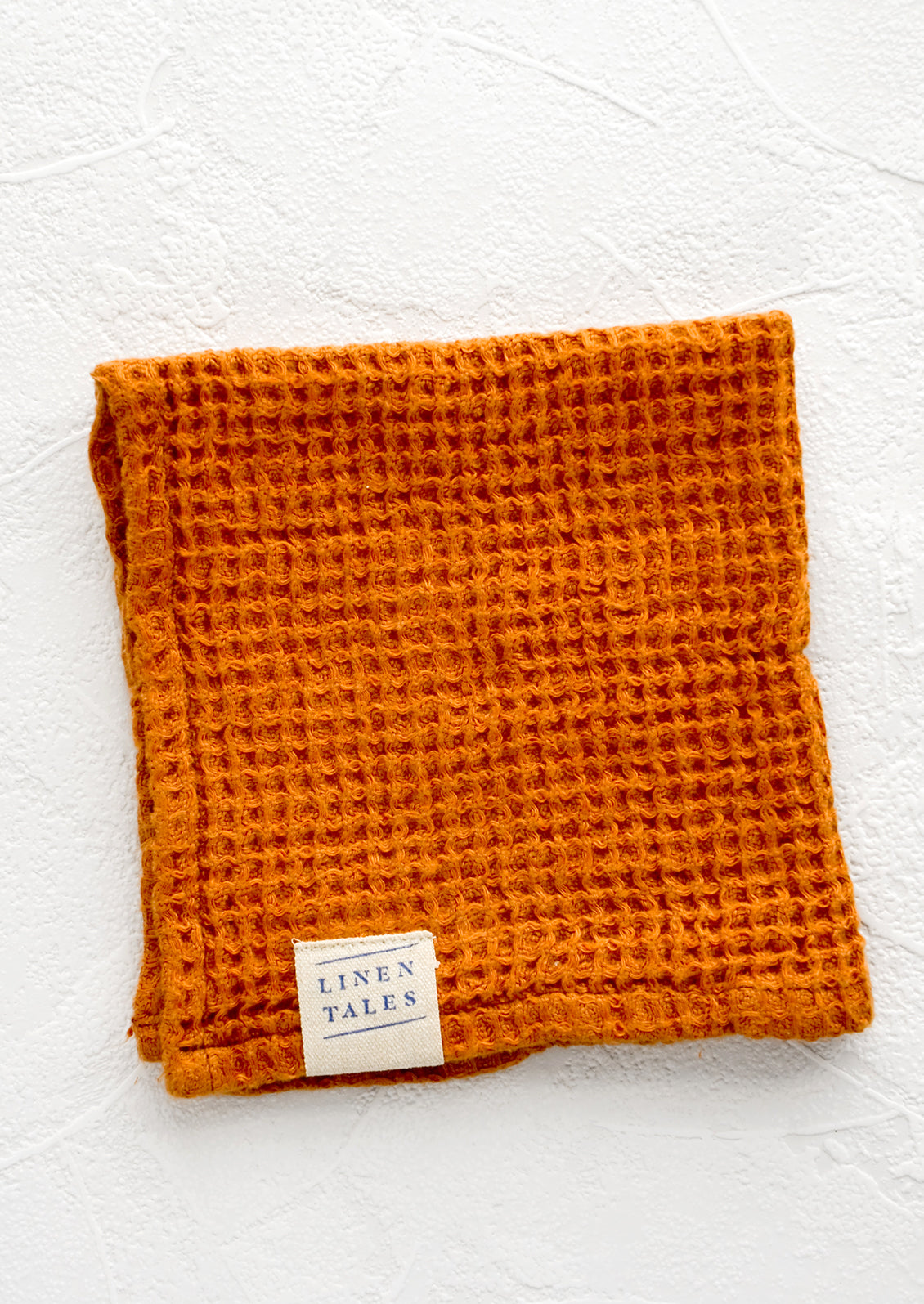 A folded waffle-weave linen washcloth in terracotta color with a logo patch.