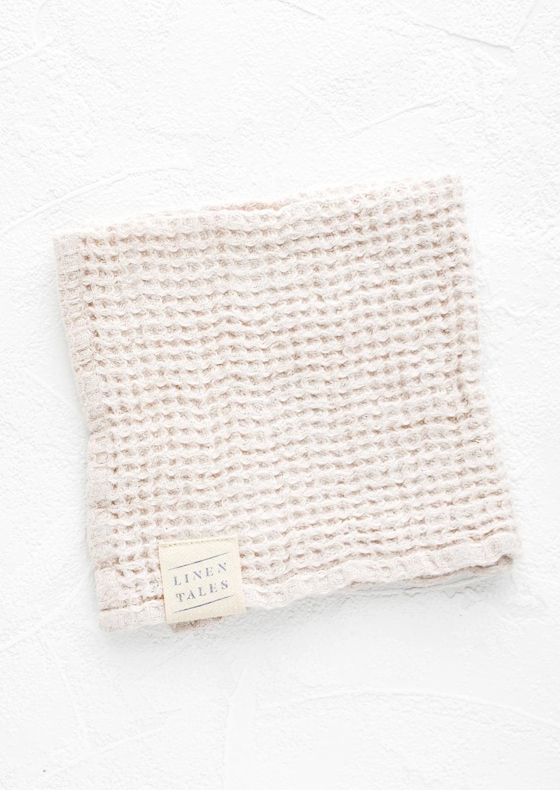 A folded waffle-weave linen washcloth in cream color with a logo patch.