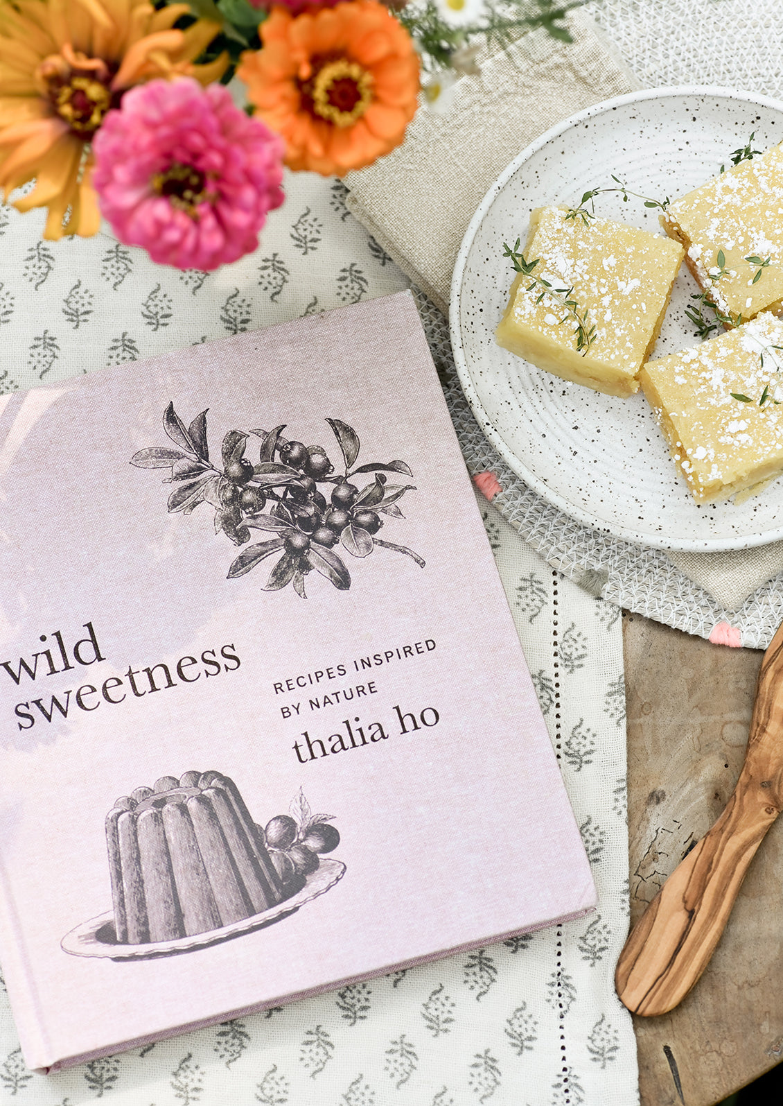 A cookbook pictured with flowers and lemon bars.